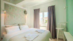 Un lit bien fait, avec deux serviettes, une chaise blanche et une vue sur l’extérieur par une fenêtre ornée de rideaux.