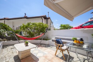 A patio with a hammock, a glass table with fruit, and a white building in the background.