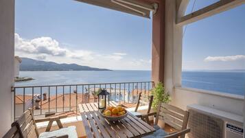 Un balcon avec une table, des chaises et un saladier rempli de fruits. La vue comprend une étendue d’eau et des terres lointaines.