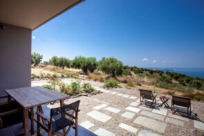 Un patio con una mesa y sillas, un sendero de piedra y vistas al mar y a los olivos.