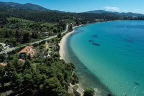 An aerial view of a coastal area with a clear beach, dense forest, and a road running parallel to the shore.