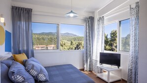 Un dormitorio con cama, televisor sobre un mueble y ventanas grandes con cortinas.