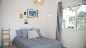 Quarto com cama, almofadas, uma cadeira de madeira, janela com vista e obras de arte nas paredes.