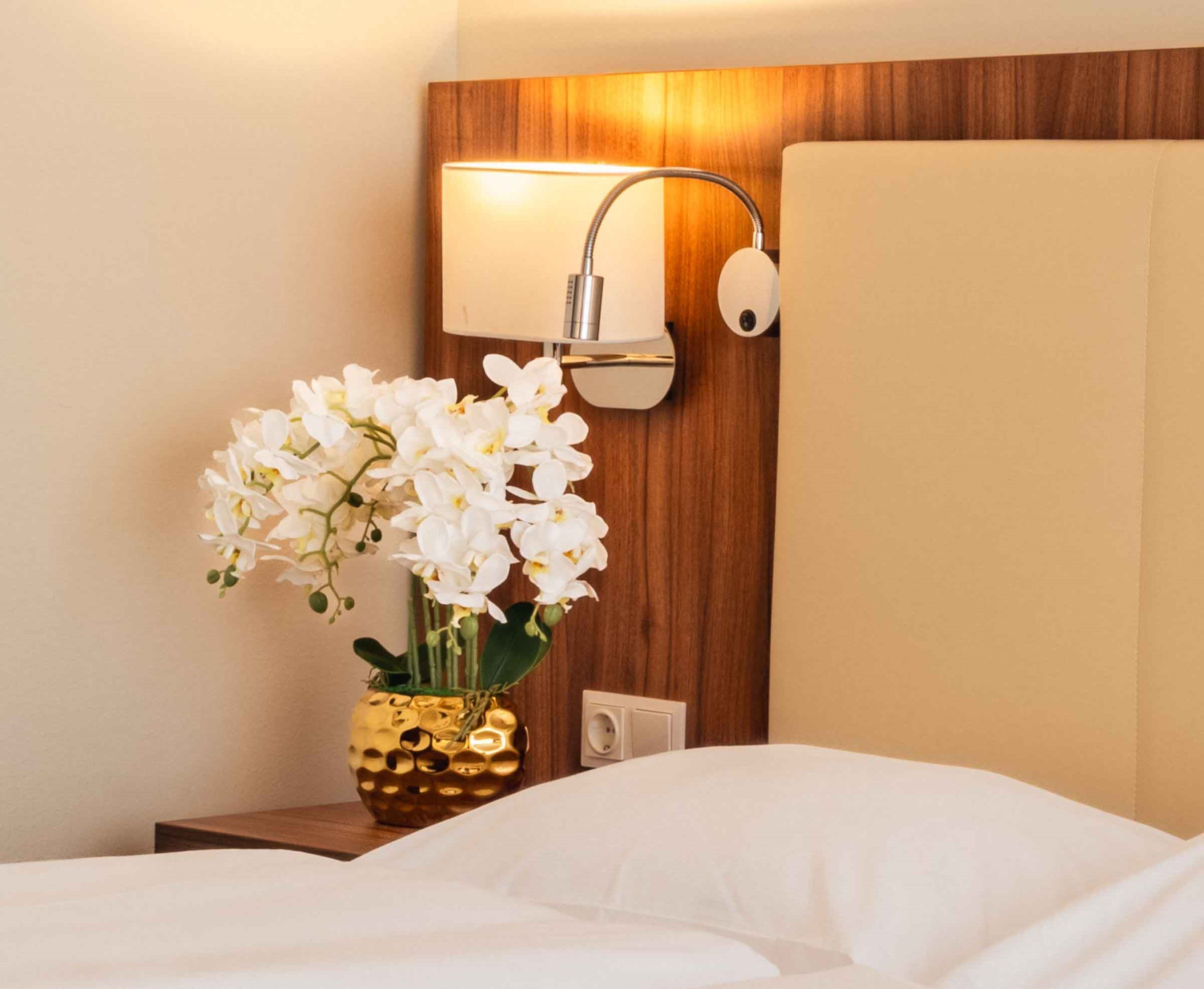 Een hotelkamer met een bed, een nachtkastje met een vaas witte bloemen, een wandlamp en een houten wandpaneel.
