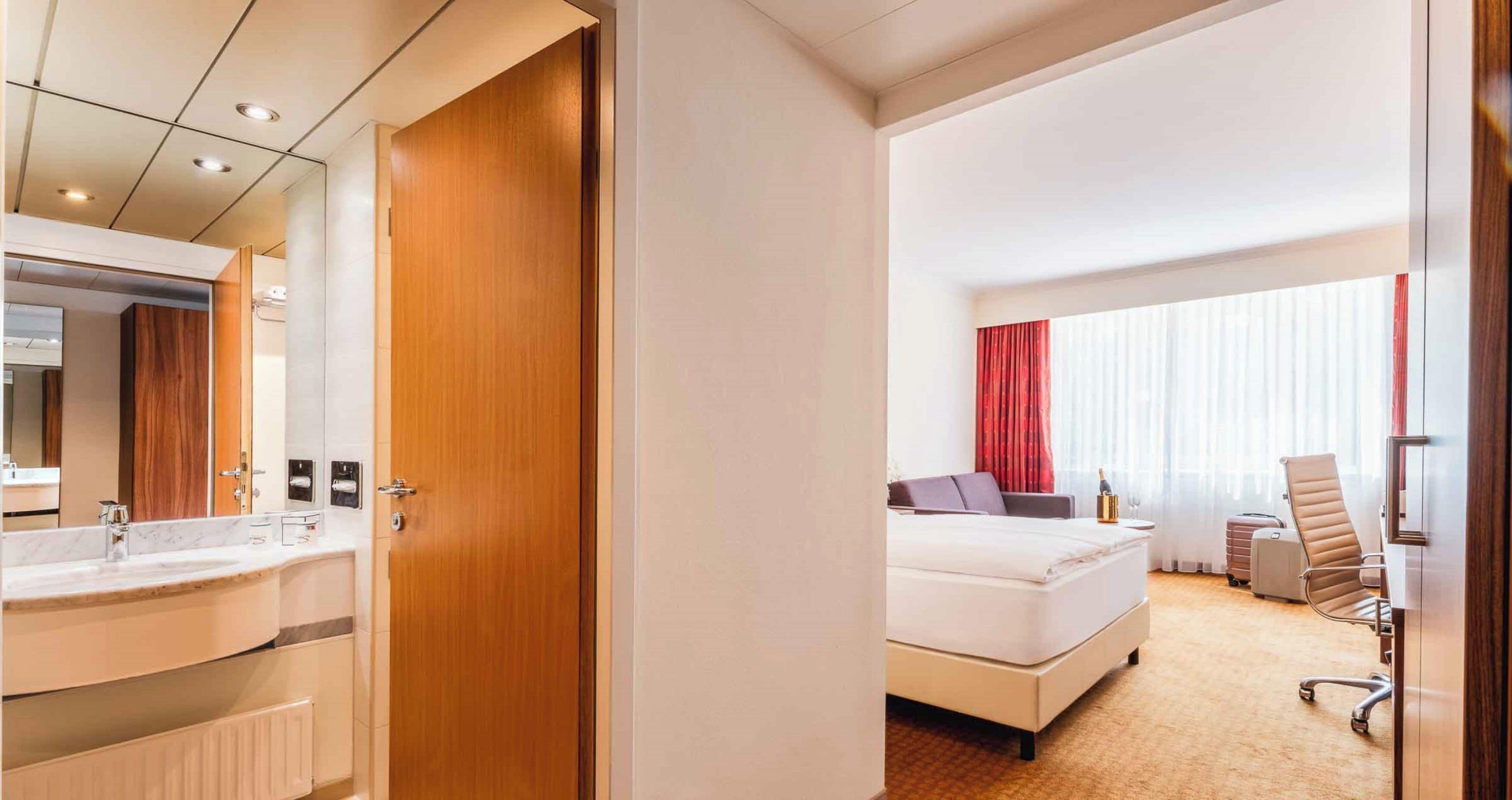 A hotel room with a wooden door, a white bed, a desk, a chair, and a bathroom with a mirror and sink.