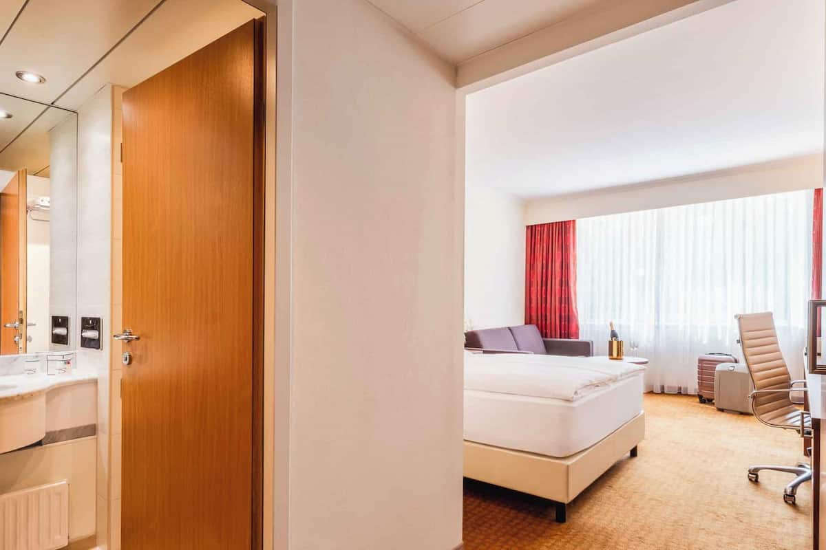 A hotel room with a wooden door, a white bed, a desk, a chair, and a bathroom with a mirror and sink.