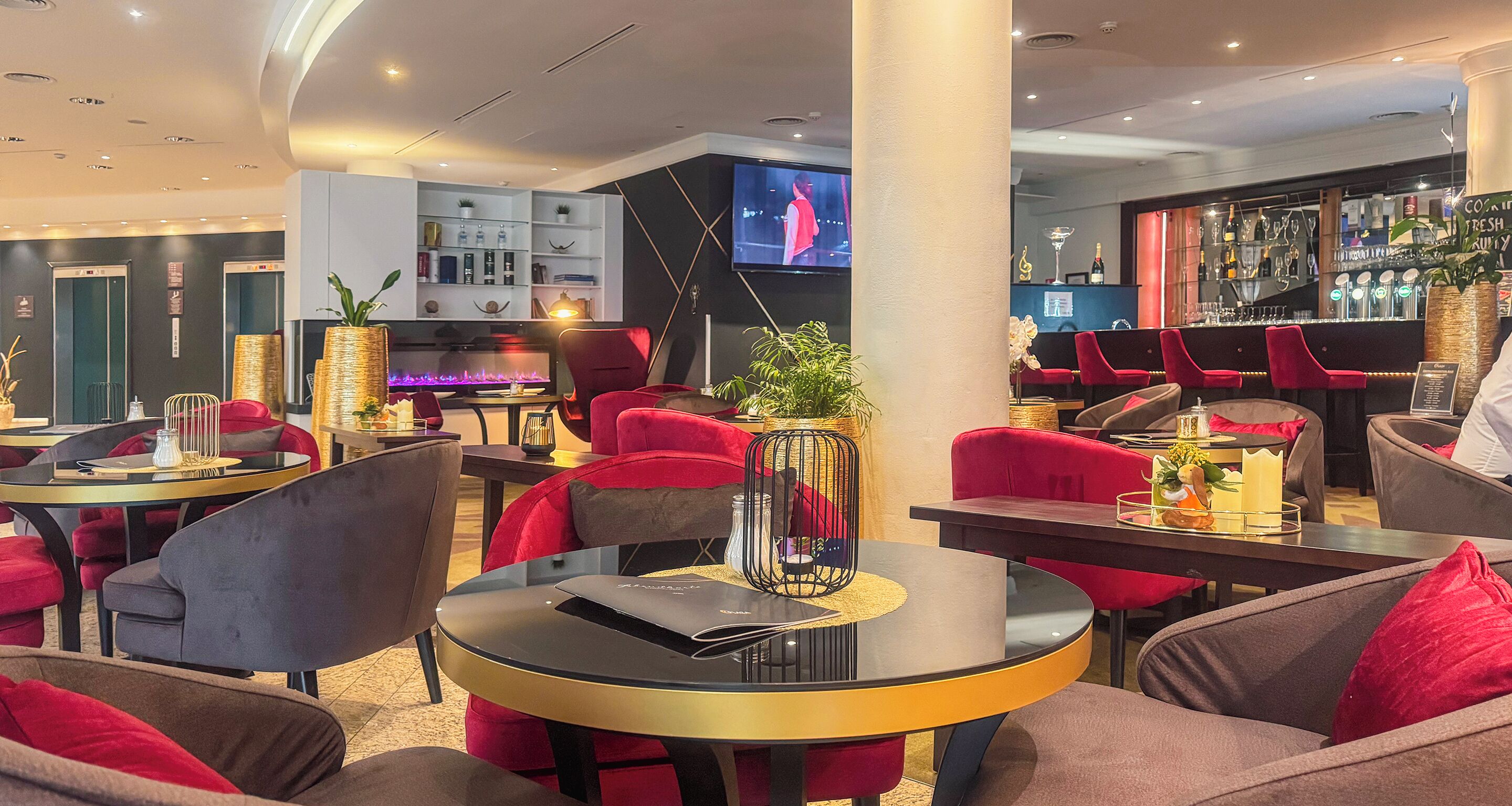 Un moderno espacio lounge con mesas redondas, sillas tapizadas en rojo y gris, un área de bar con sillas rojas y un televisor montado en la pared.