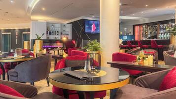 Un moderno espacio lounge con mesas redondas, sillas tapizadas en rojo y gris, un área de bar con sillas rojas y un televisor montado en la pared.