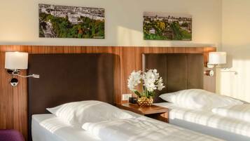 Habitación de hotel con dos camas, cabeceros de madera, ropa de cama blanca, lámparas fijadas a la pared y dos cuadros enmarcados con vistas a la ciudad.