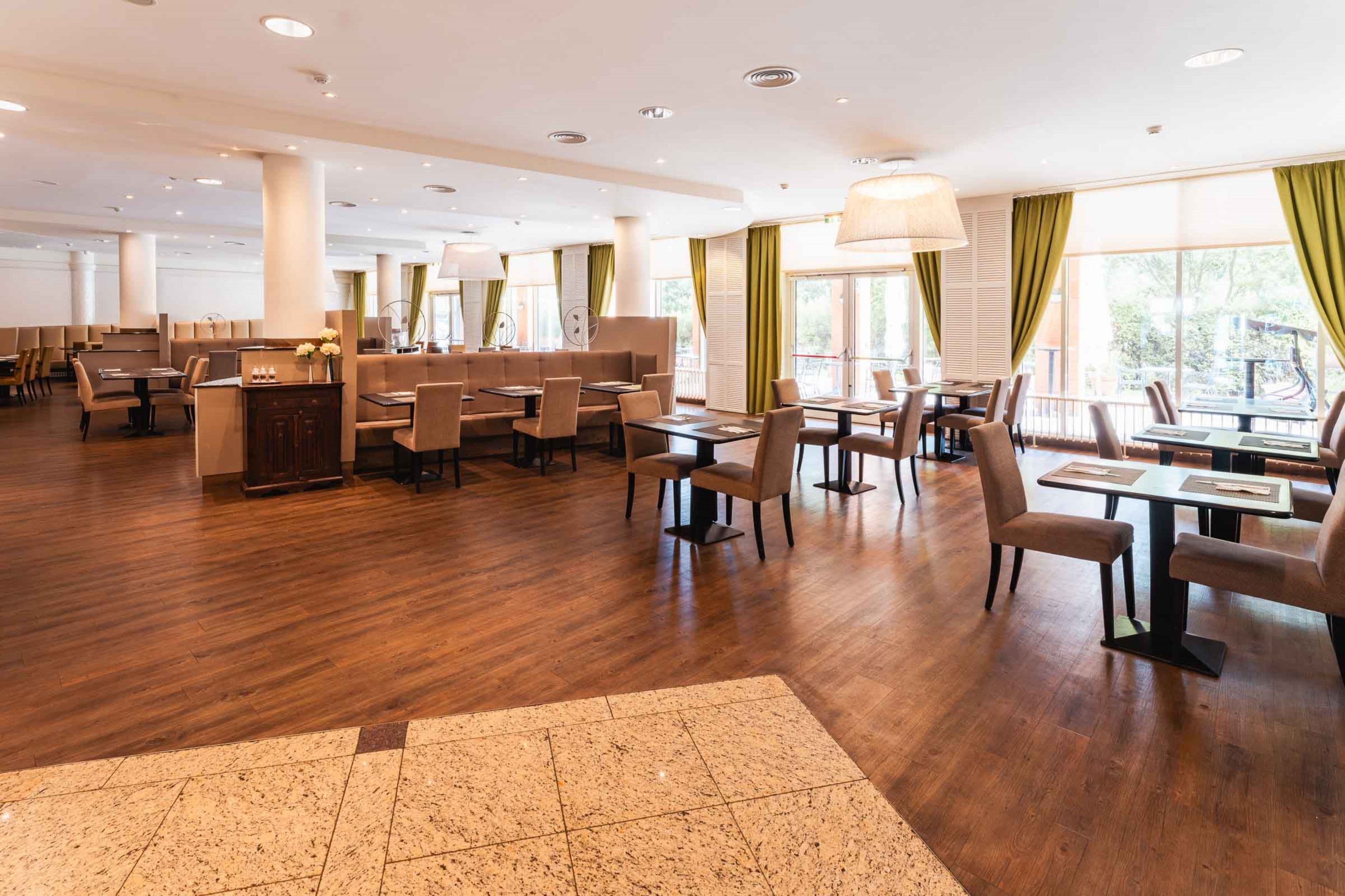 Spaziosa sala da pranzo di un hotel, con pavimento in legno, diversi tipi di disposizione dei posti a sedere e ampie finestre con tende verdi.