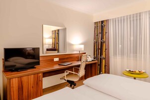 Quarto de hotel com uma escrivaninha de madeira, televisão, cadeira, cama, mesinha com frutas e uma janela grande com cortinas leves.