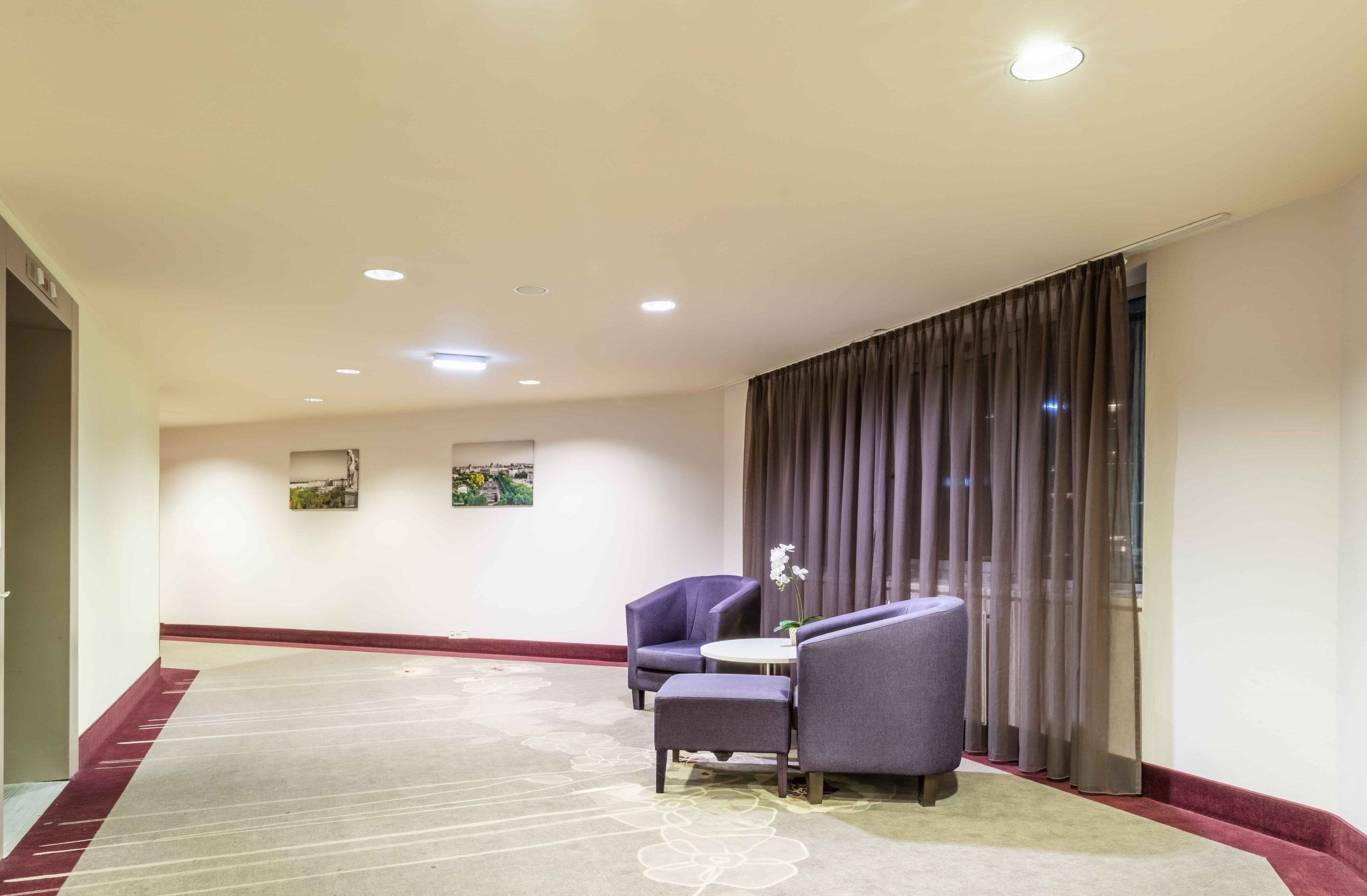 Un vestíbulo de hotel con piso alfombrado, dos sillones, una mesita y un amplio ventanal con cortinas.