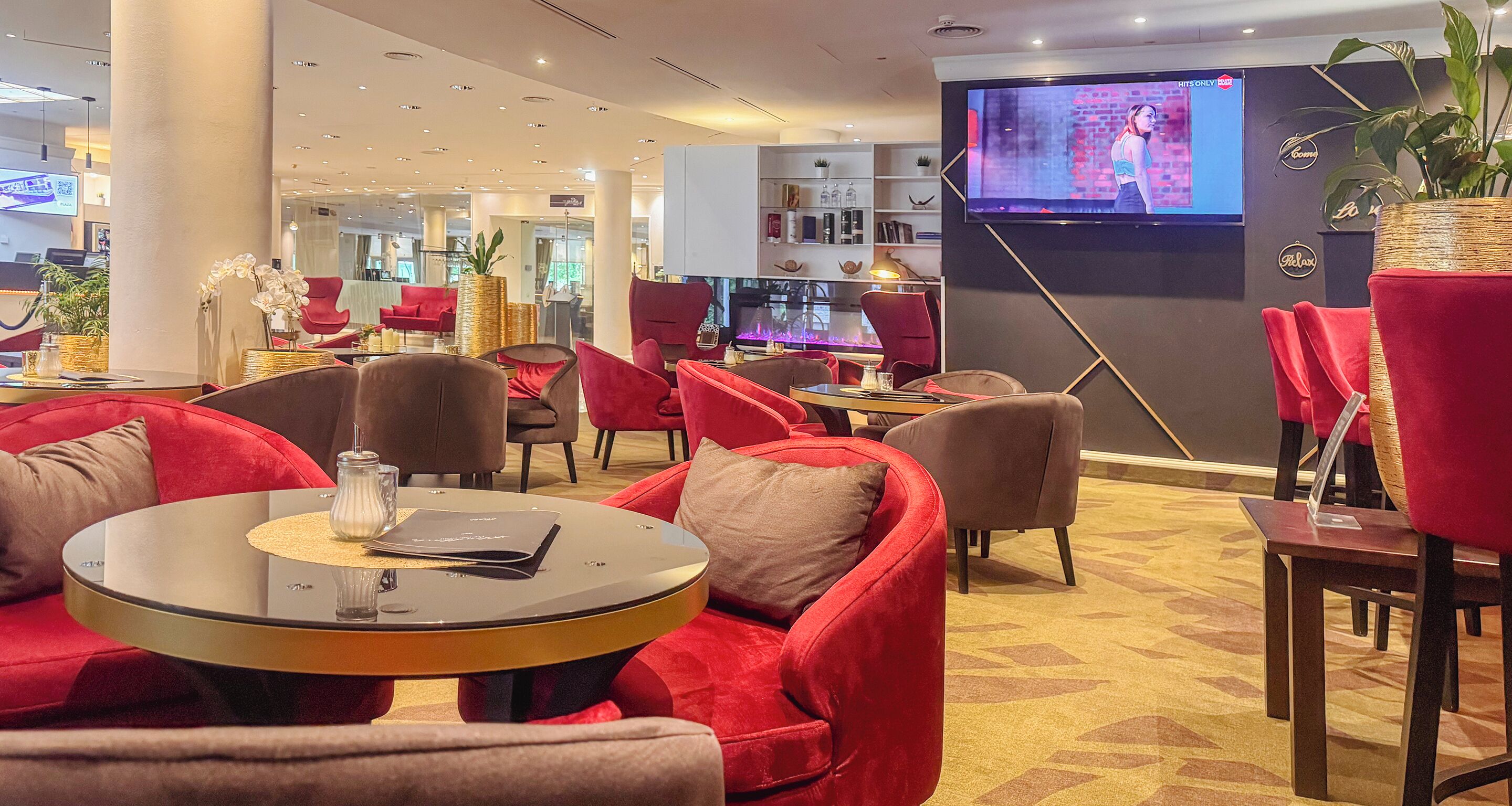 A modern hotel lobby with red upholstered seating, a round glass table, and a large flat-screen TV mounted on the wall.
