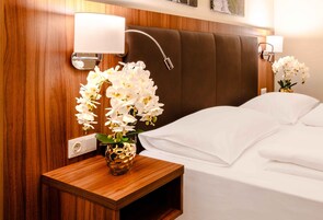 Camera d'albergo con una testiera in legno, un comodino con un vaso di fiori bianchi e due lampade a parete.