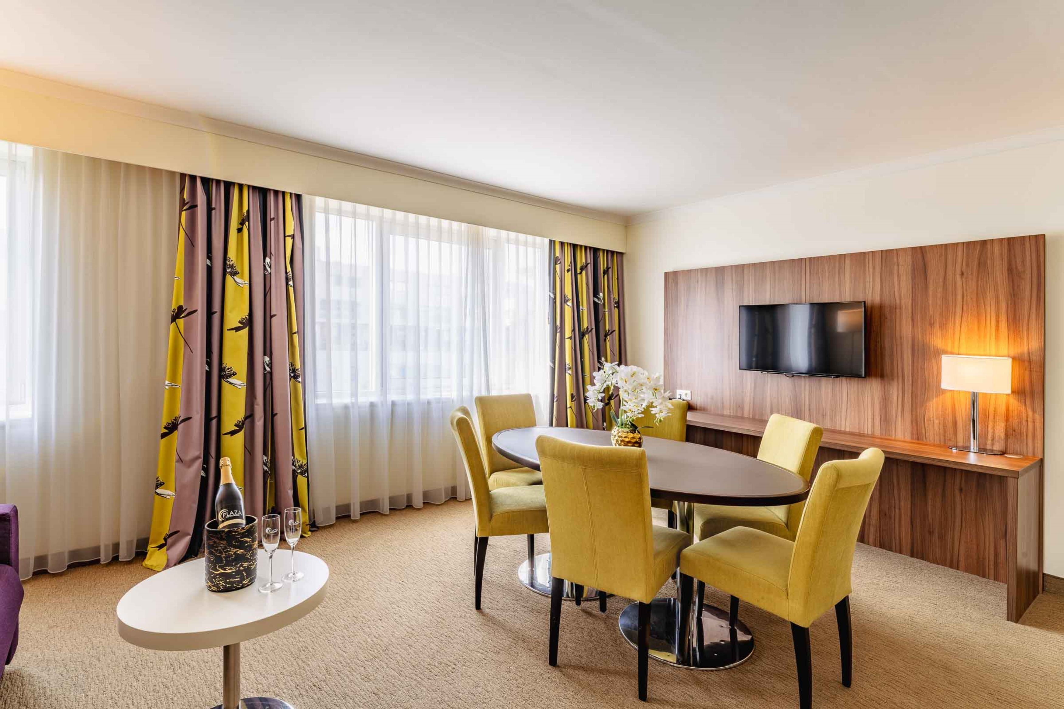 Ein Hotelzimmer mit Essbereich, ausgestattet mit einem runden Tisch, gelben Stühlen und einer Champagnerflasche. Ein Flachbildfernseher und eine Lampe sind an der Wand befestigt.