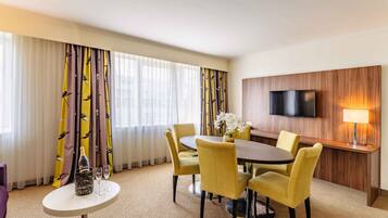 Ein Hotelzimmer mit Essbereich, ausgestattet mit einem runden Tisch, gelben Stühlen und einer Champagnerflasche. Ein Flachbildfernseher und eine Lampe sind an der Wand befestigt.