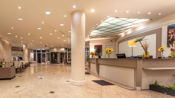 A hotel lobby with a reception desk, a glass door entrance, a seating area with chairs, and a large pillar. - PLAZA Premium Wien (Vienna)