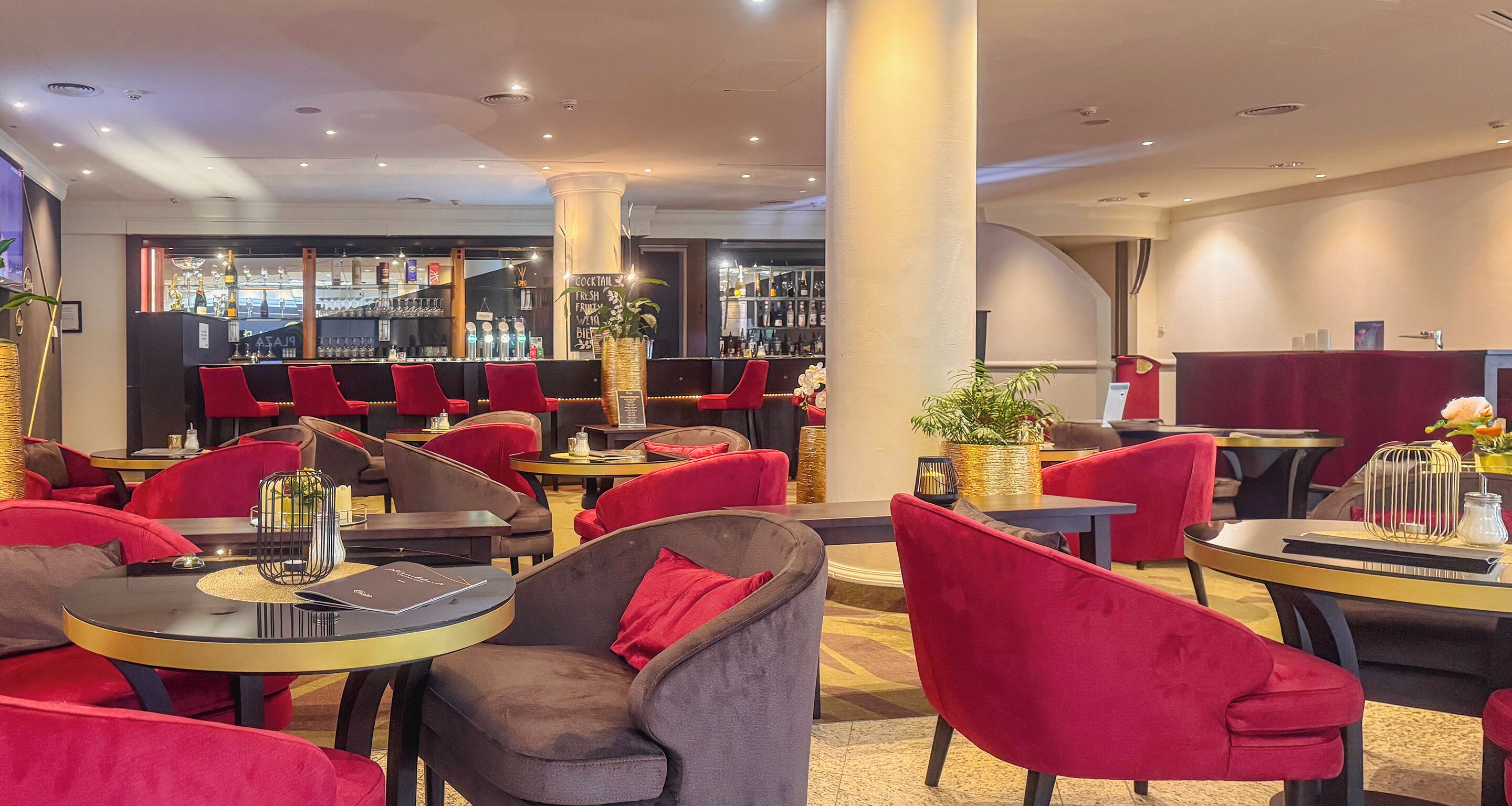 Un interior de restaurante moderno con sillas tapizadas en rojo y gris, mesas redondas y una zona de bar al fondo.