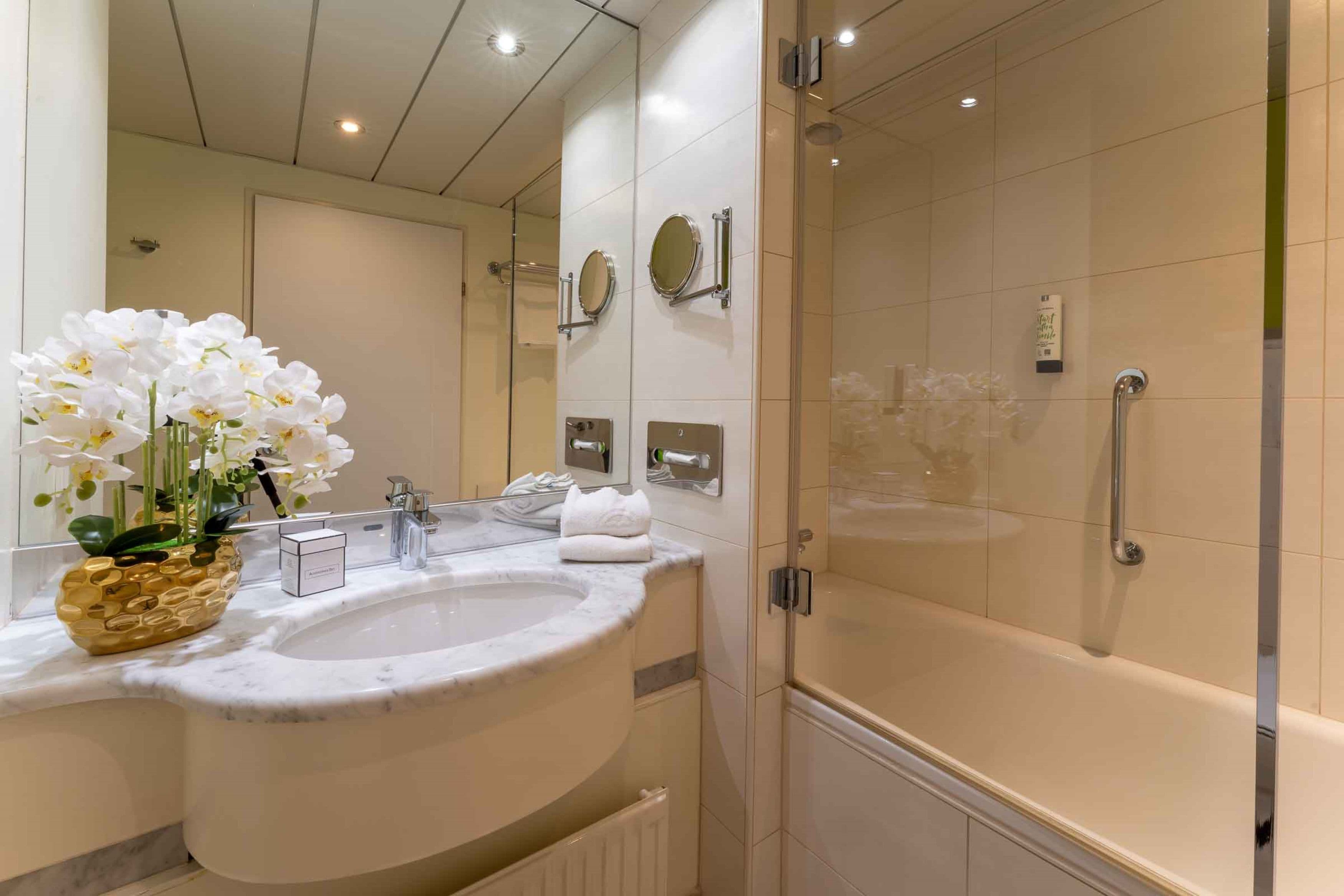 Ein modernes Badezimmer mit Marmorwaschbecken, zwei Spiegeln, einer Duschkabine und einer dekorativen Orchideenpflanze.