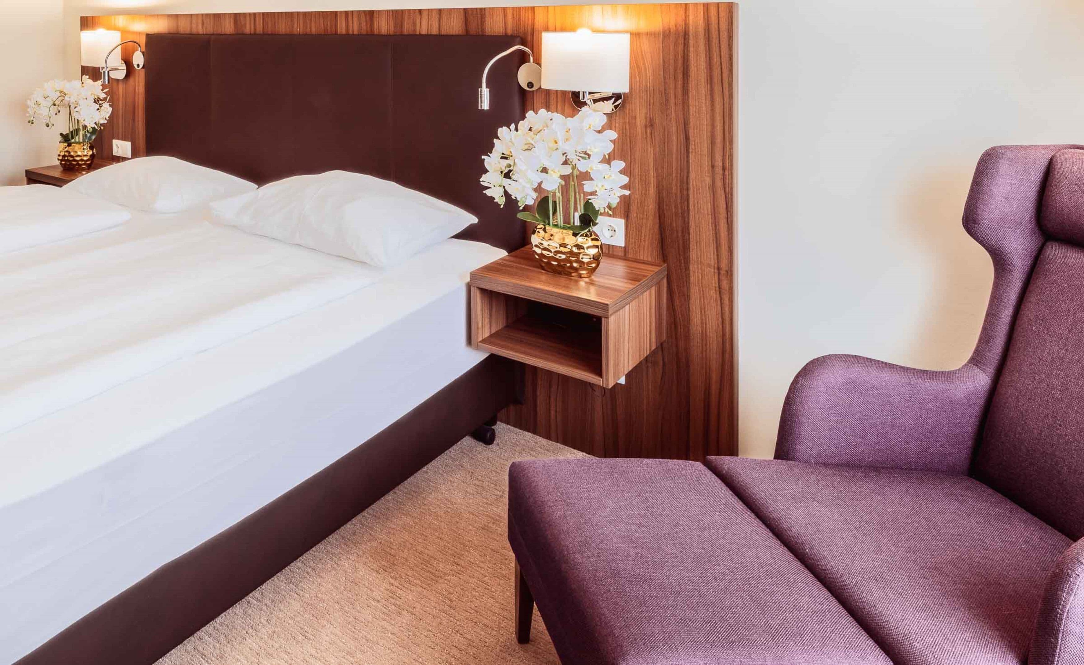 Quarto de hotel com uma cama grande, uma mesinha de cabeceira com abajur, uma pequena mesa com flores e uma poltrona roxa.