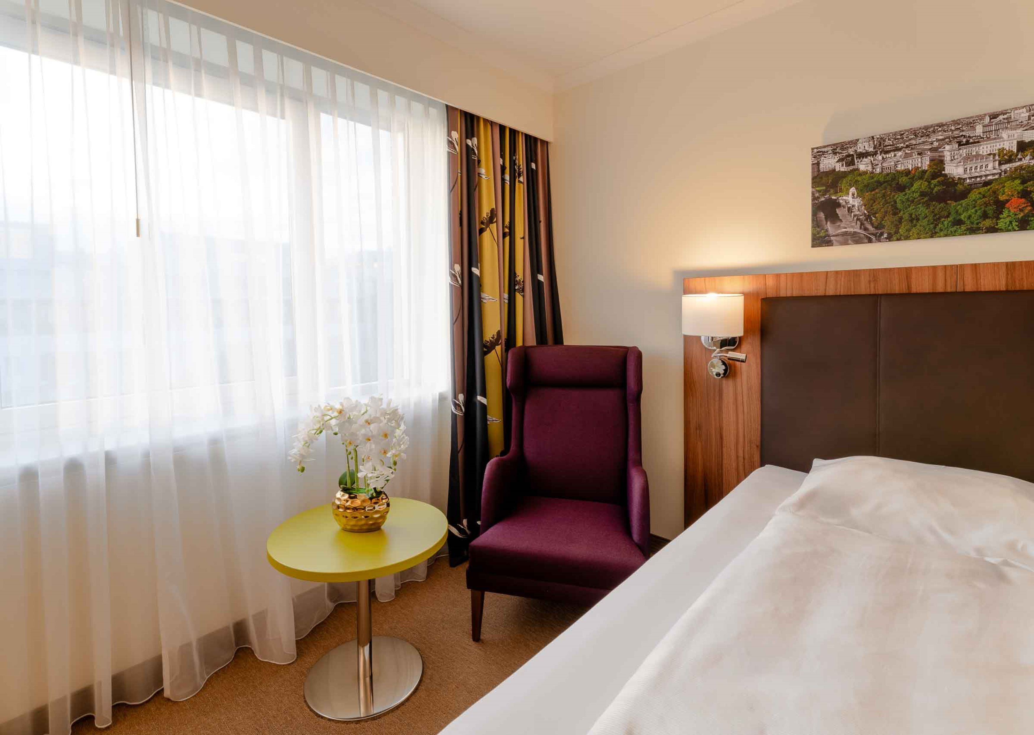 Ein Hotelzimmer mit einem Bett, einem violetten Sessel, einem kleinen runden Tisch mit Blumenvase und Blick auf die Stadt durch das Fenster.
