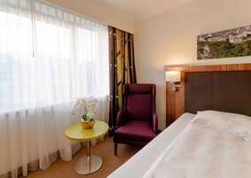 Ein Hotelzimmer mit einem Bett, einem violetten Sessel, einem kleinen runden Tisch mit Blumenvase und Blick auf die Stadt durch das Fenster.