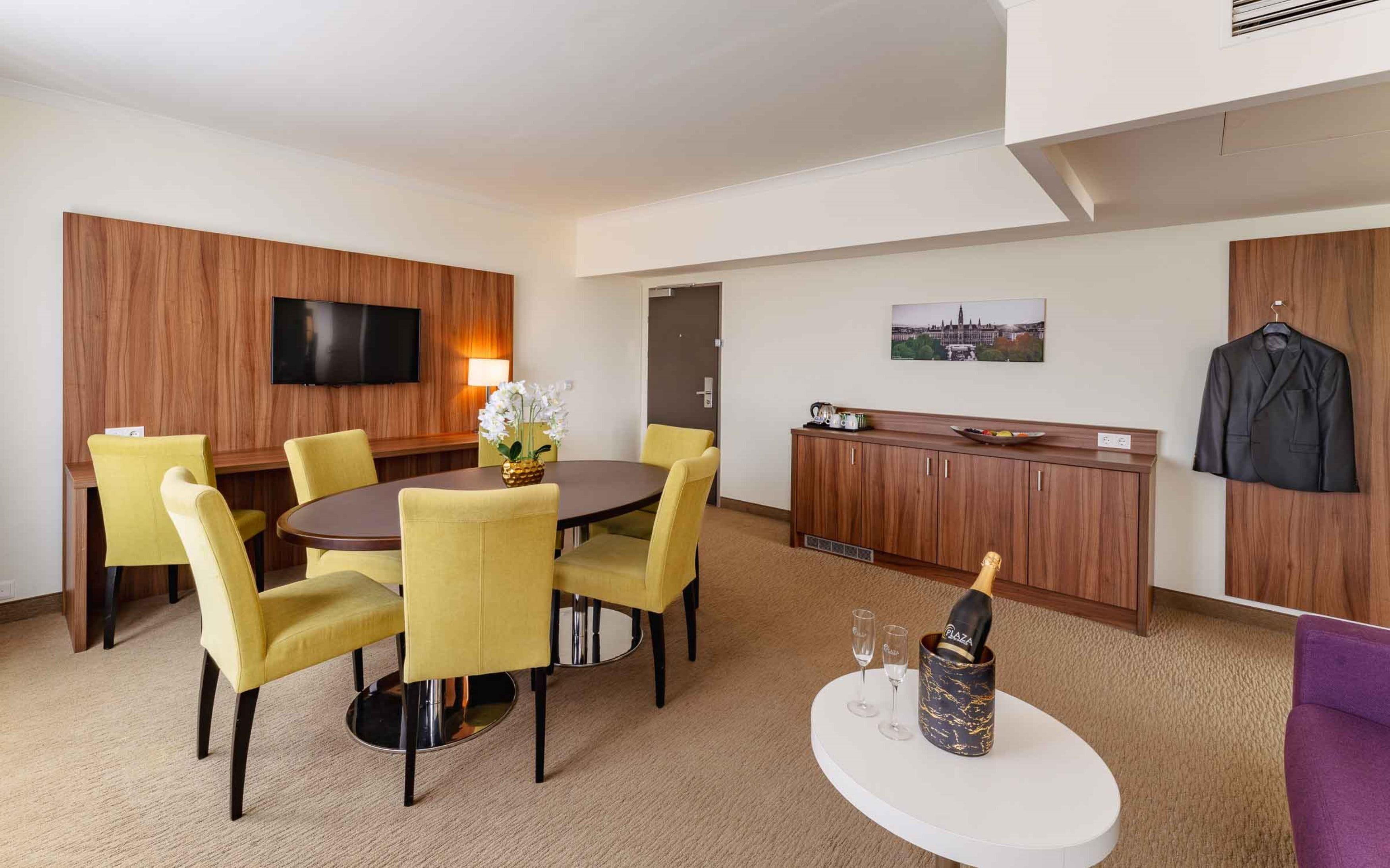 A hotel room with a round dining table, yellow chairs, a flat-screen TV, a minibar, a coat rack, and a cityscape painting.
