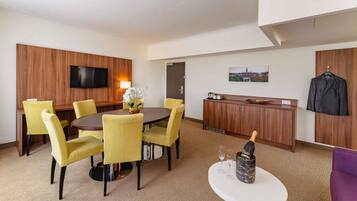 A hotel room with a round dining table, yellow chairs, a flat-screen TV, a minibar, a coat rack, and a cityscape painting.