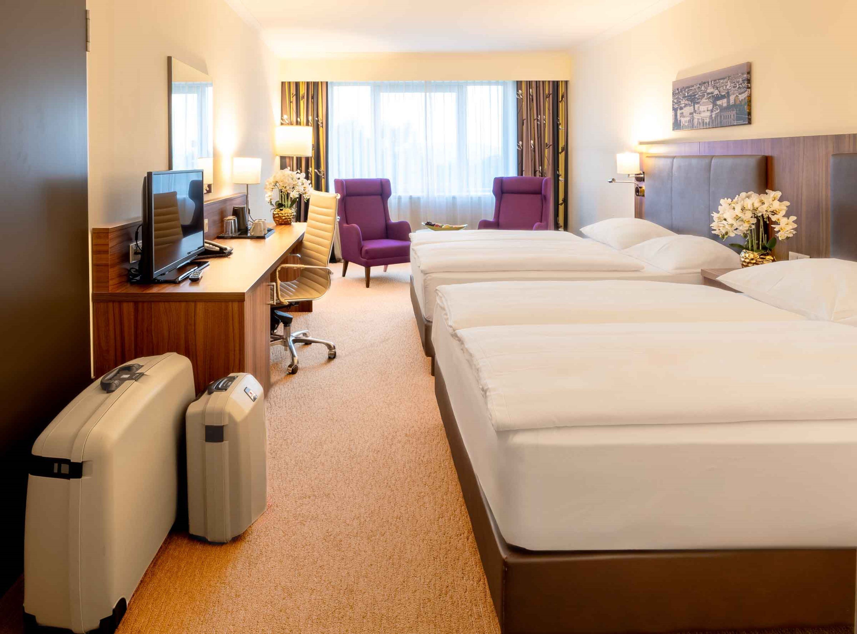 Une chambre d’hôtel avec un bureau, deux fauteuils violets, un lit et un téléviseur.