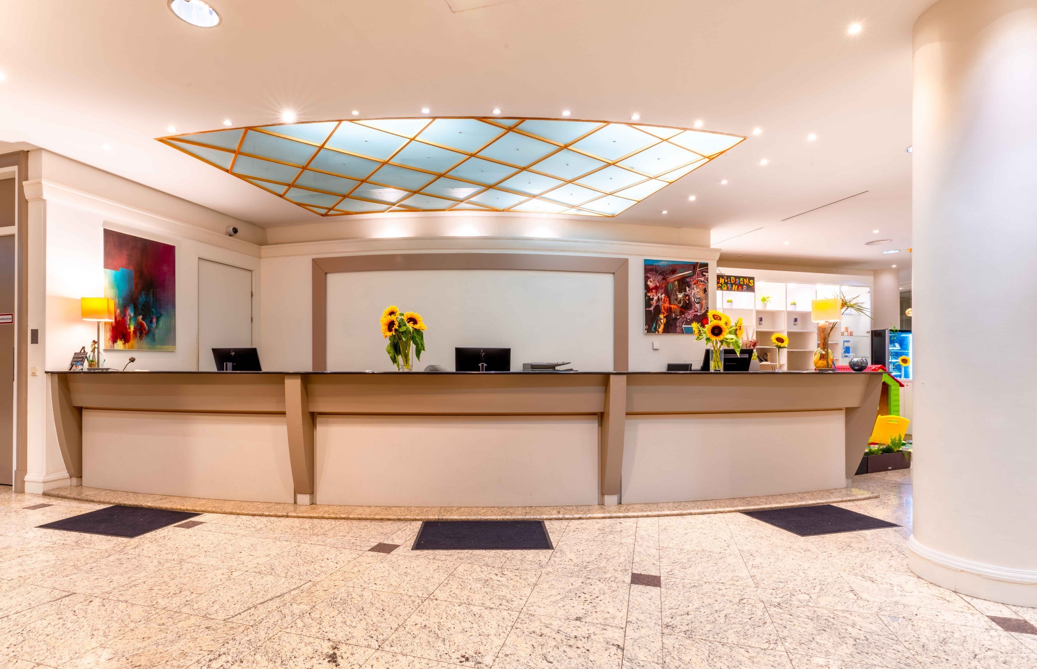 A hotel reception area with a modern design, featuring a central desk, a ceiling light fixture, and a wall-mounted artwork.