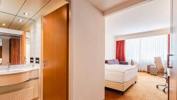 A hotel room with a wooden door, a white bed, a desk, a chair, and a bathroom with a mirror and sink.