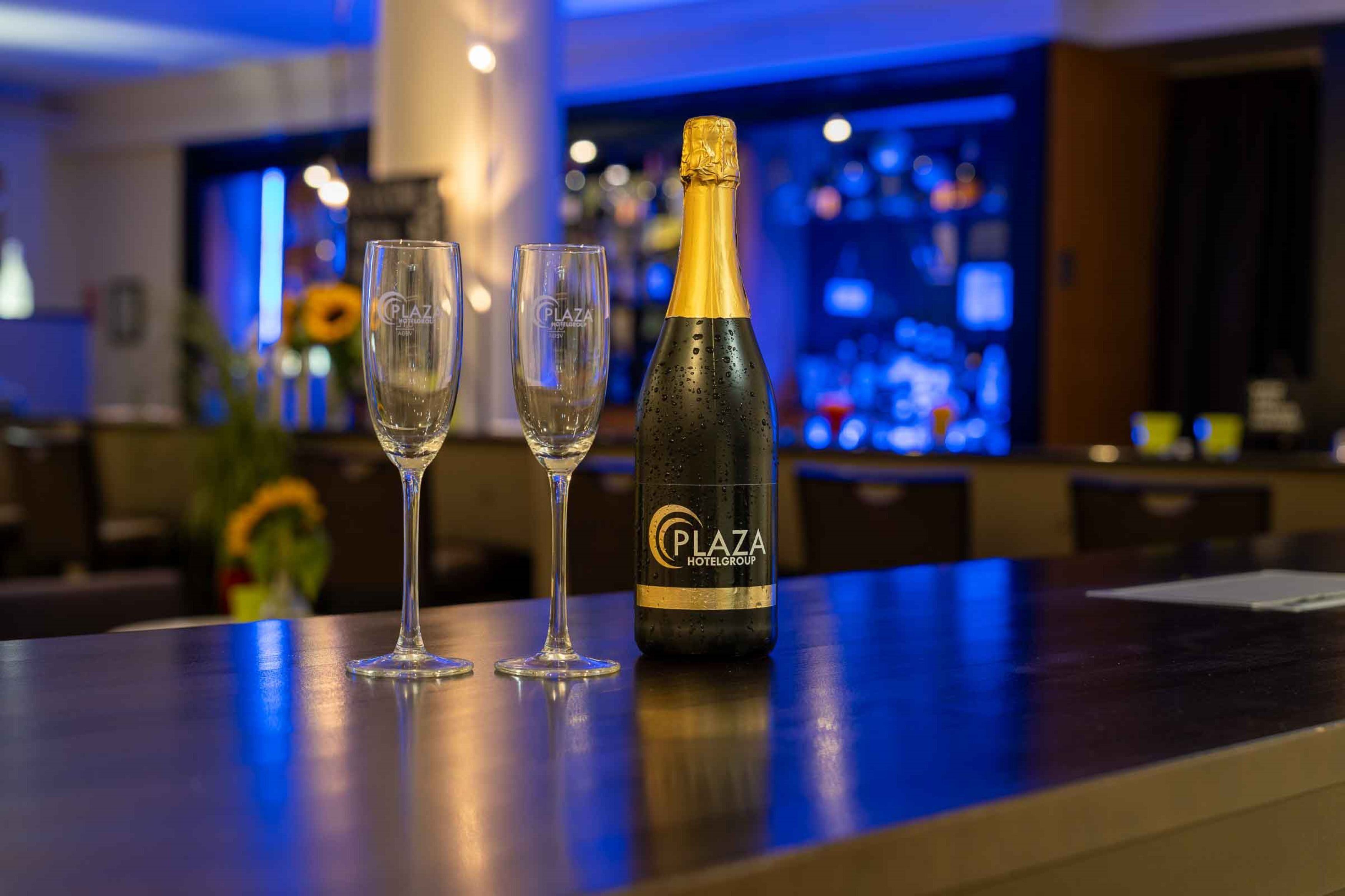 Un'area bar con una bottiglia di champagne Plaza Hotelgroup e due bicchieri vuoti su un bancone.