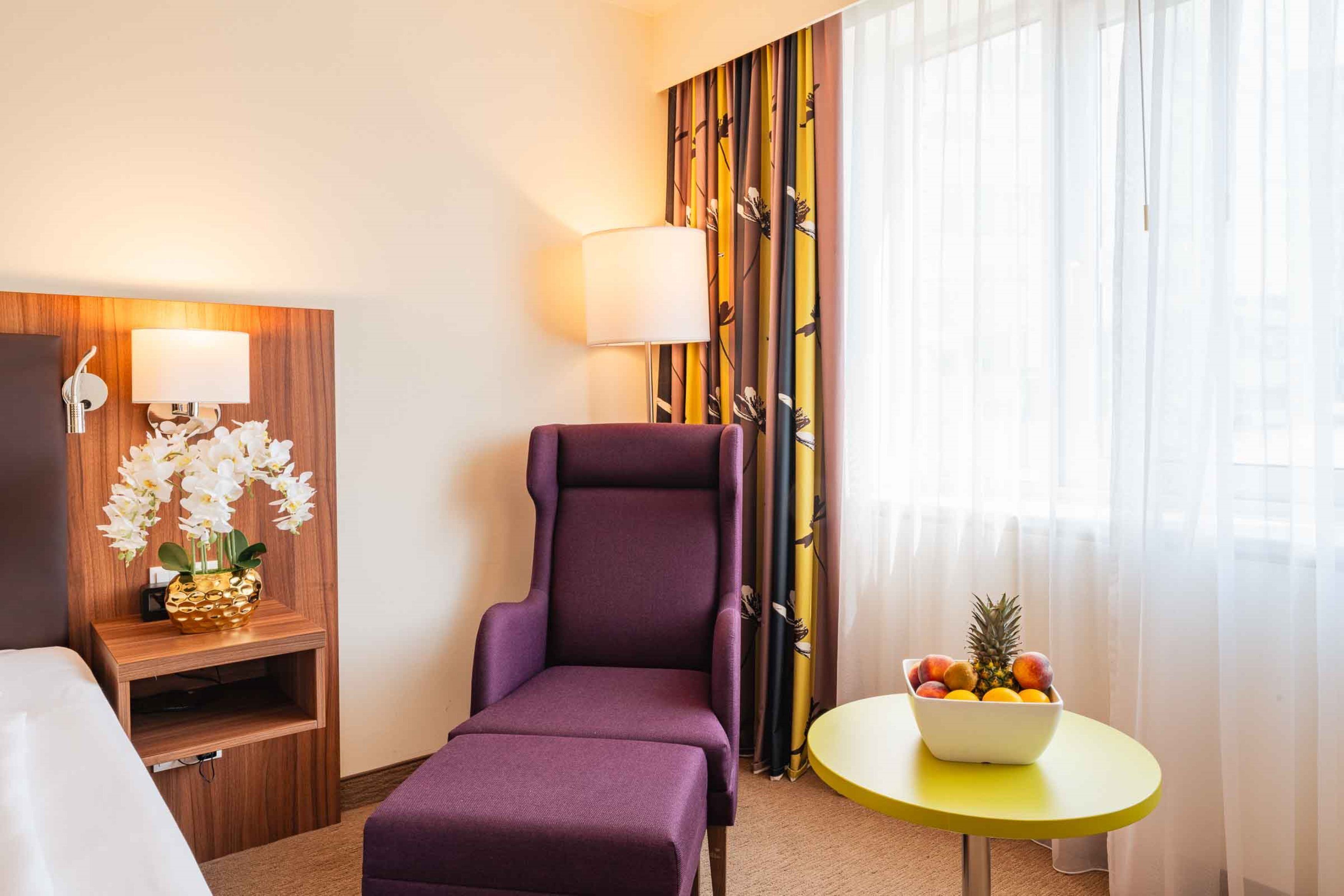 Quarto de hotel com uma poltrona roxa, uma mesa pequena com uma fruteira, uma televisão sobre um suporte de madeira e uma luminária fixada na parede.