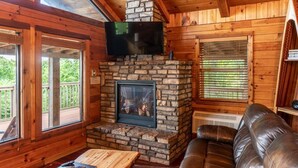 A cozy cabin interior with a stone fireplace, leather sofa, and a flat-screen TV mounted above it.