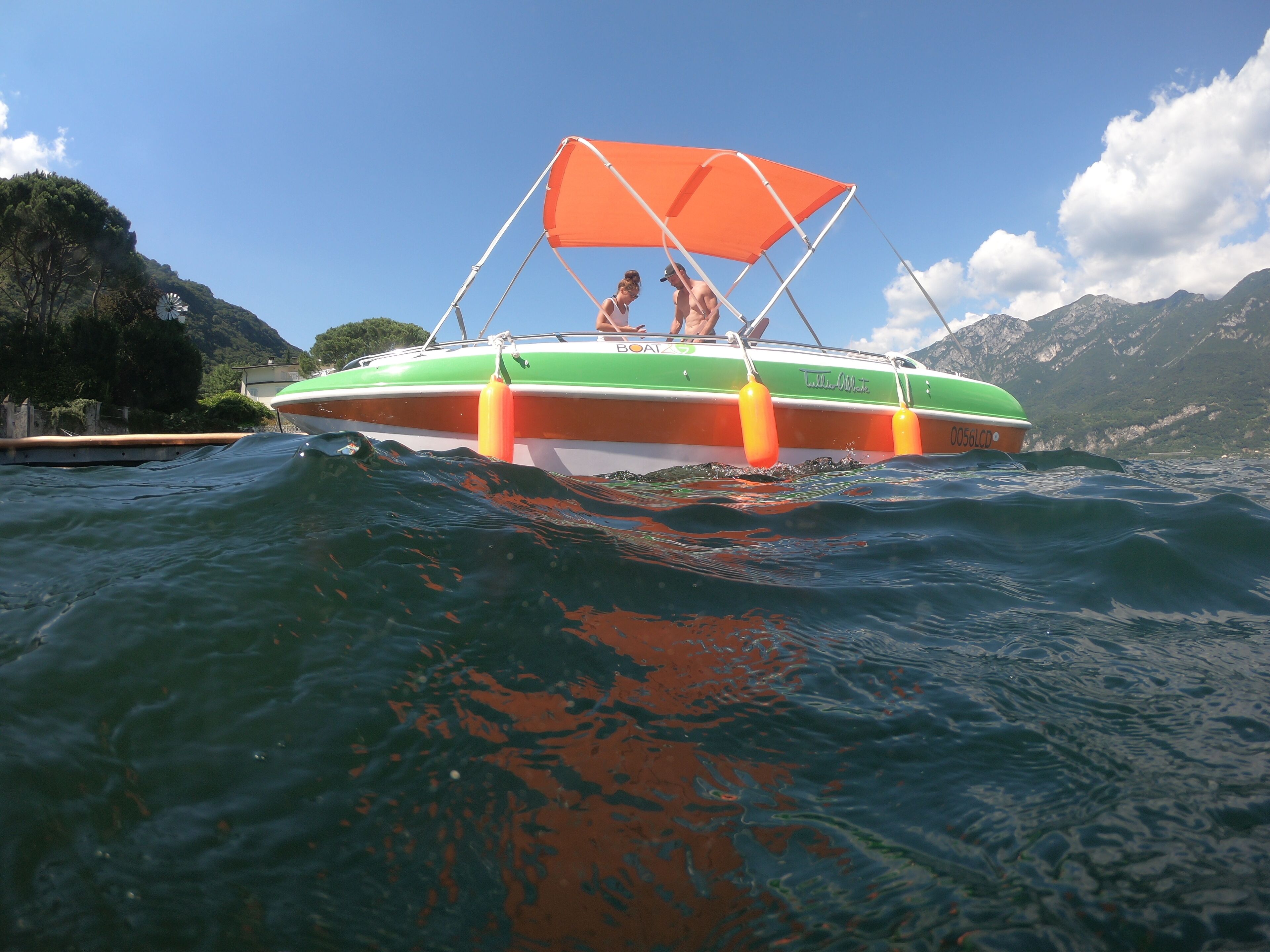 Un bateau vert et orange, surmonté d’un auvent orange, flotte sur l’eau, avec des montagnes en arrière-plan.
