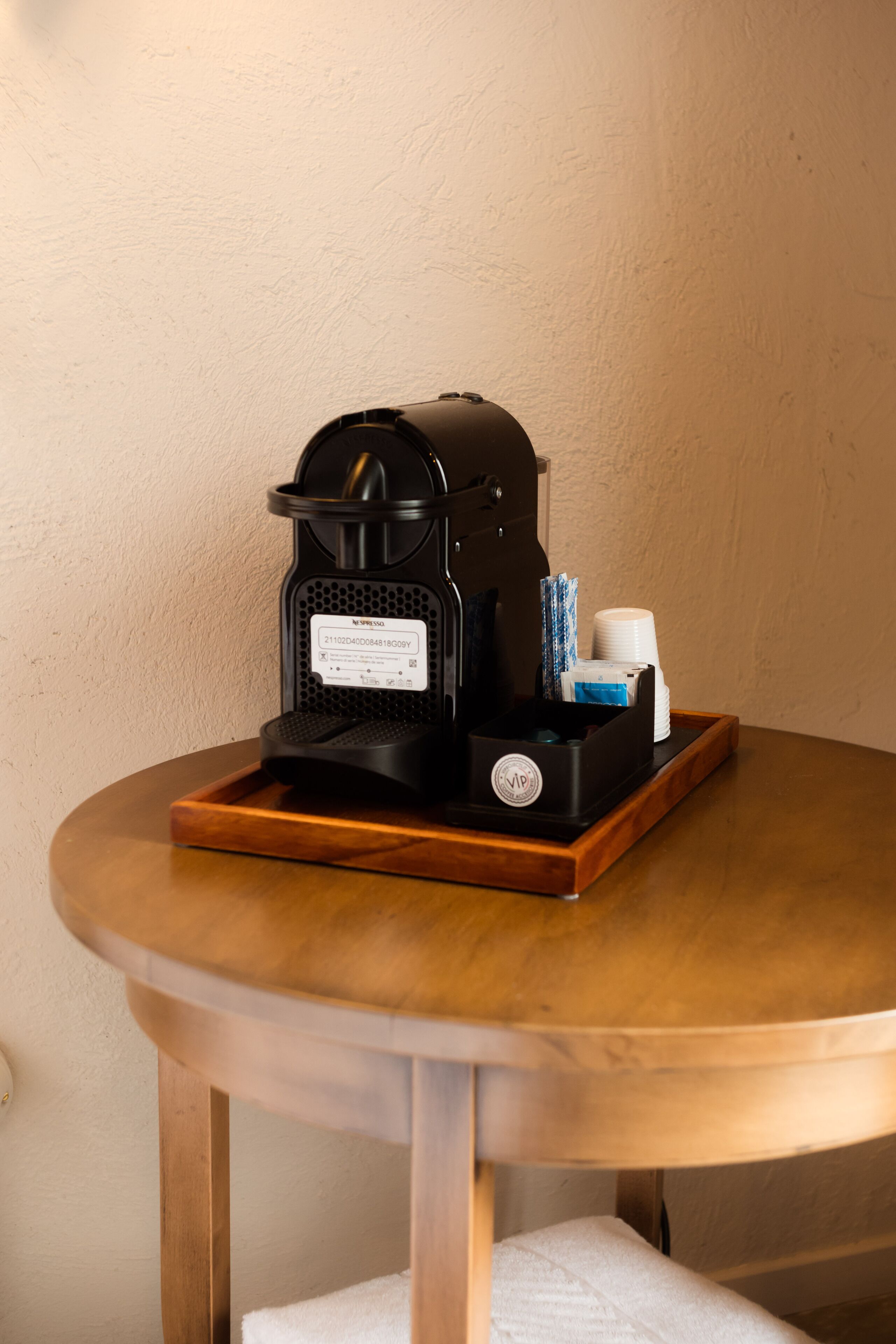 A coffee machine on a wooden table with a tray, a white wall, and a wooden chair with a white cushion.