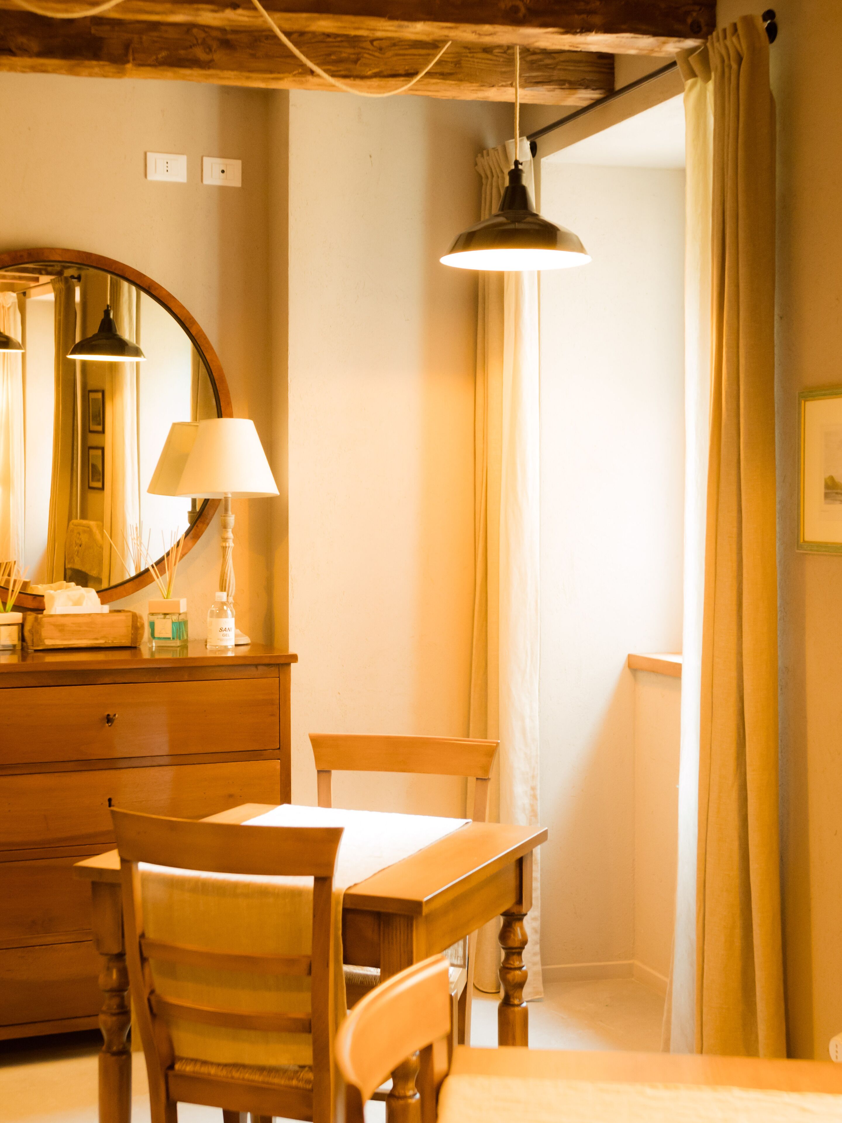 A wooden dining table with chairs, a mirror, and a lamp in a room with a wooden ceiling and a window with curtains.