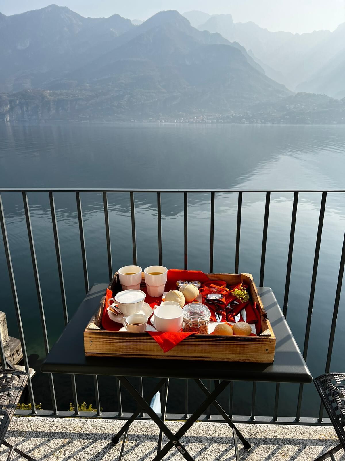 A table with a tray of food and drinks on a balcony overlooking a lake and mountains.