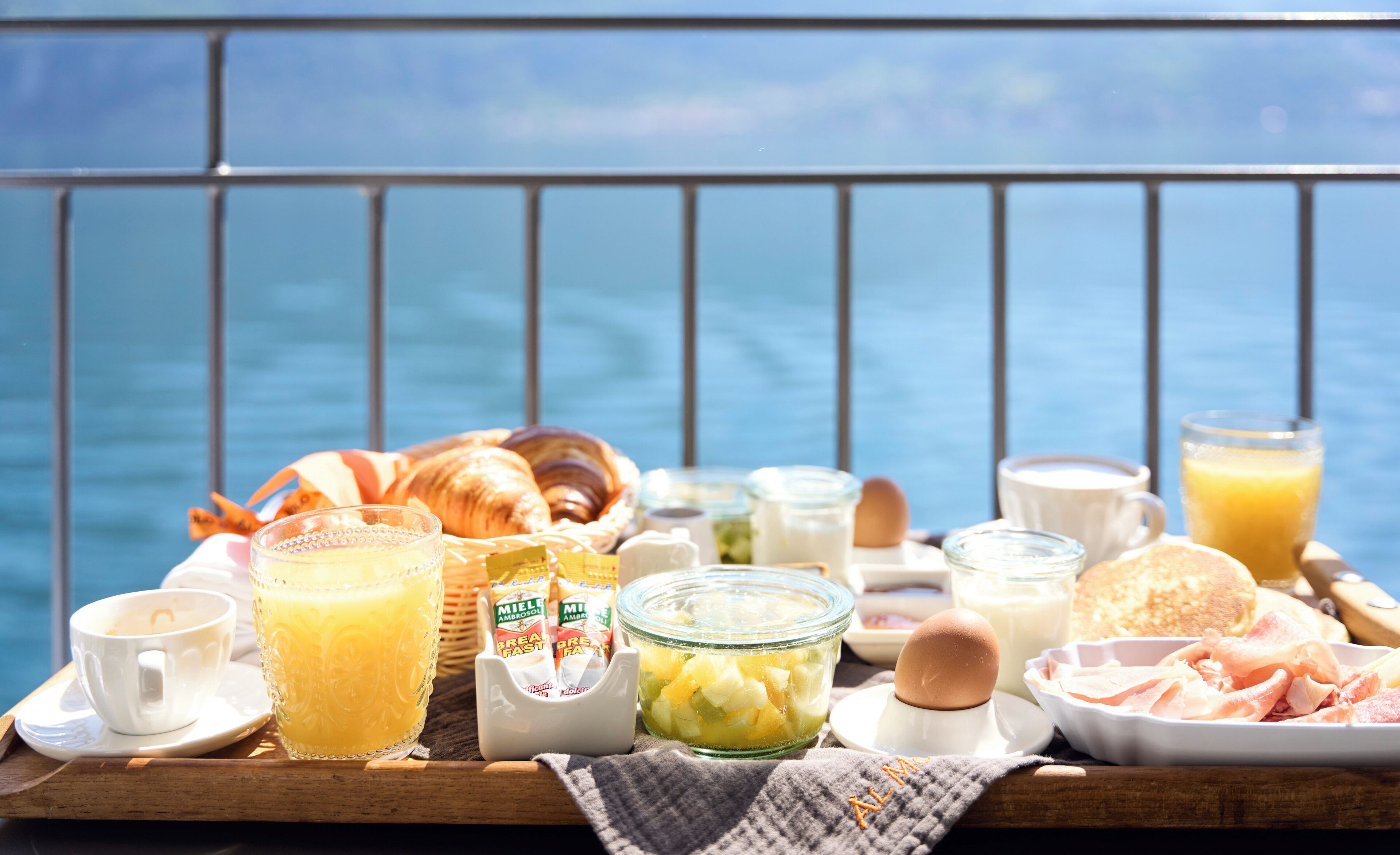 Bandeja de desayuno con cruasanes, huevos, leche y jugo en un balcón con vista al mar.