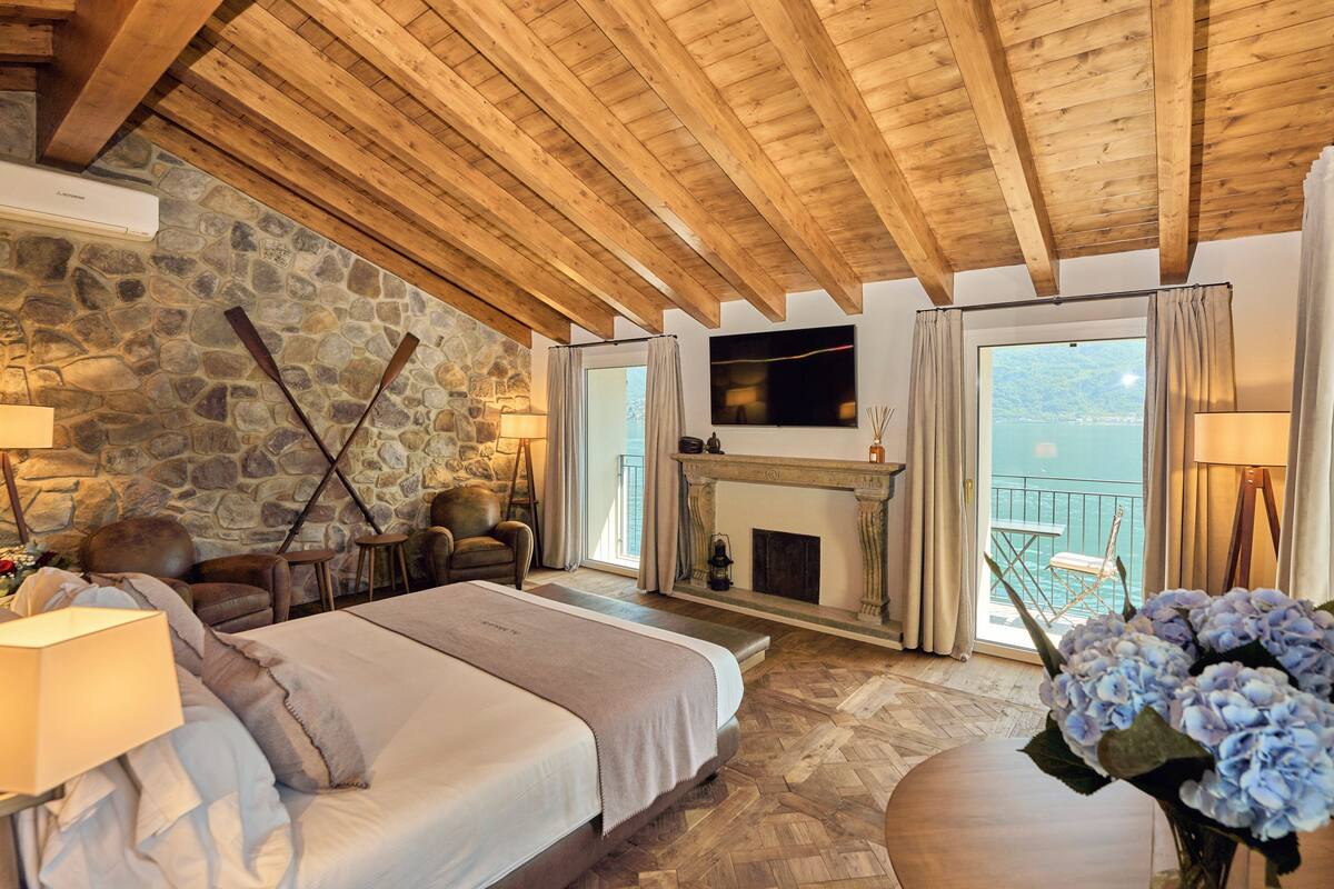 A spacious bedroom with a stone wall, wooden ceiling beams, a fireplace, and a balcony with a view of the sea.