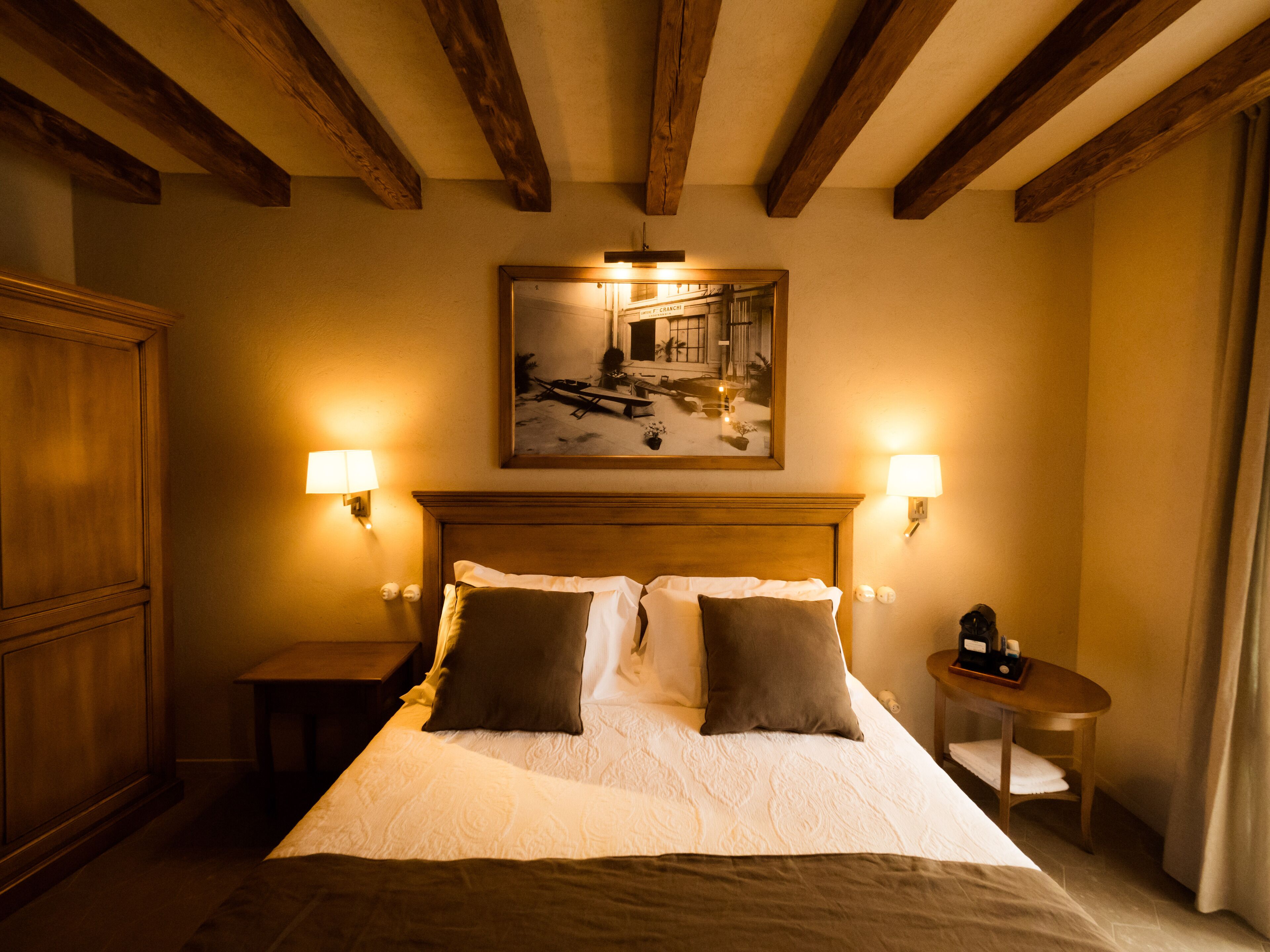 A hotel room with a large bed, wooden headboard, two bedside lamps, a nightstand, a small table with a lamp, and a painting on the wall.