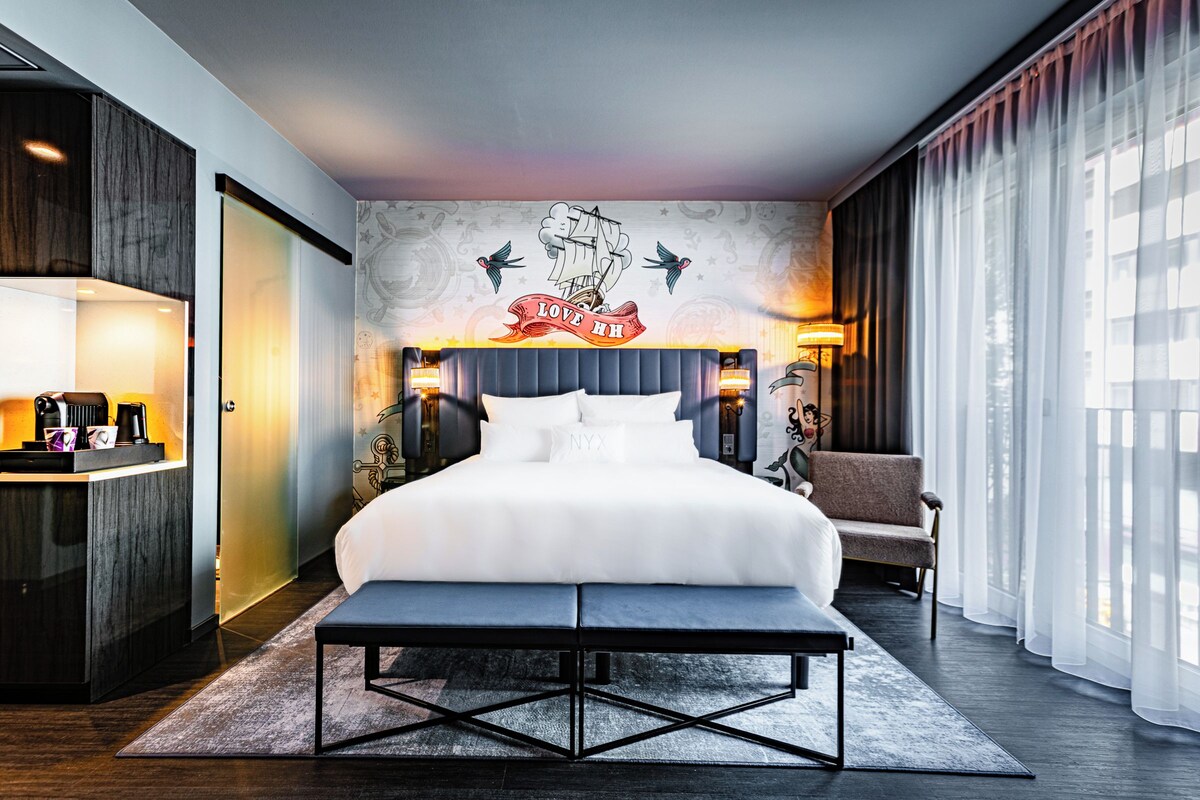 A modern hotel room with a large bed, a desk with a lamp, a chair, and a decorative wall mural.