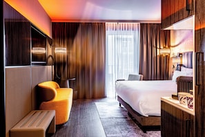 A modern hotel room with a large bed, a seating area with a sofa and ottoman, a television, and a desk with a lamp. - NYX Hotel Hamburg by Leonardo Hotels (Hamburg)