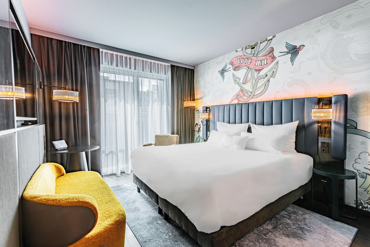 Quarto de hotel moderno com uma cama grande, um sofá, uma mesinha e um mural decorativo na parede.