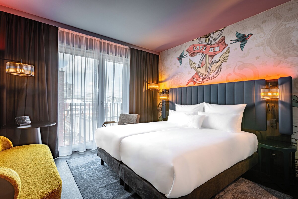 A hotel room with a large bed, a chair, a small table, and a wall mural.