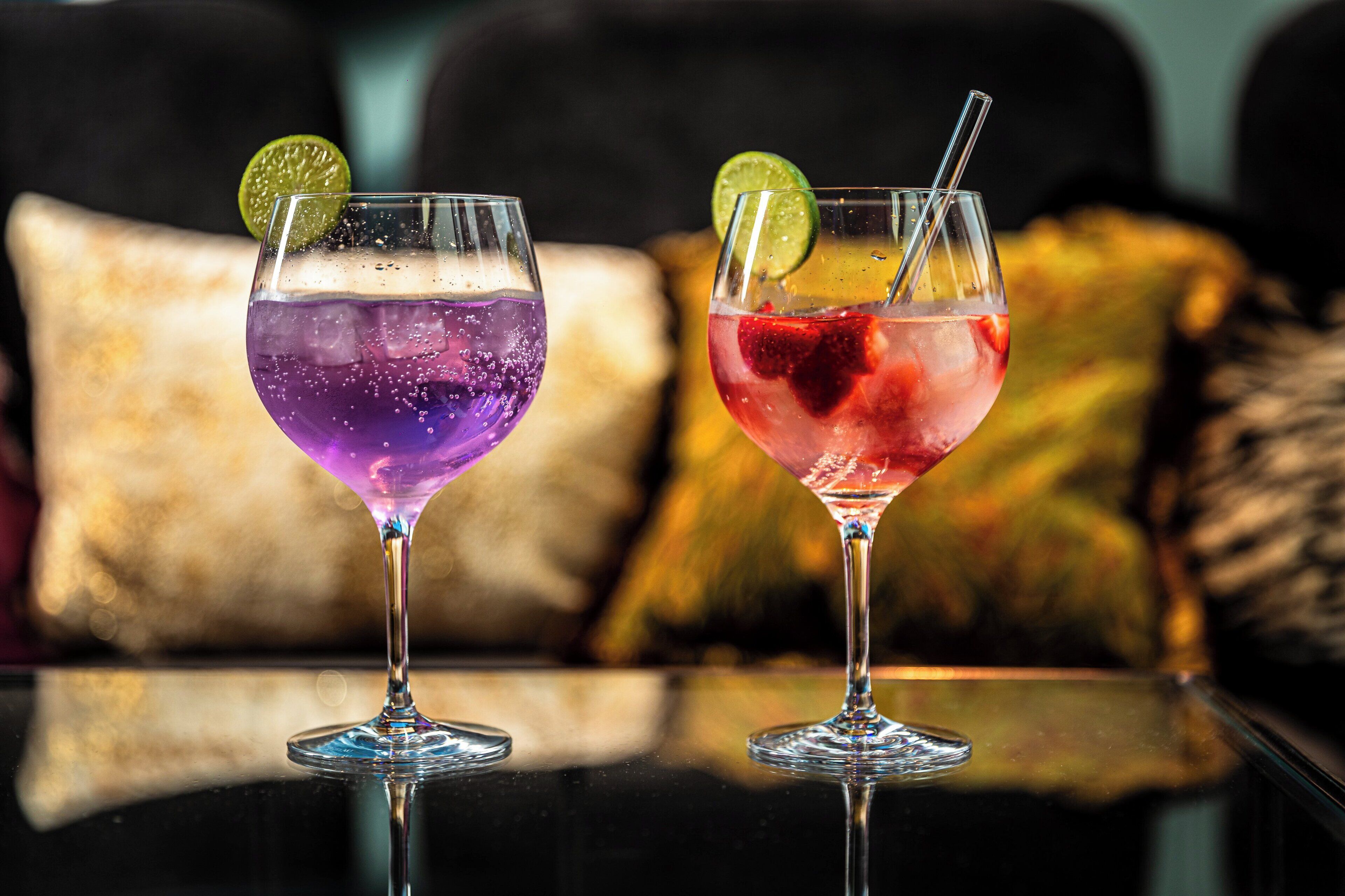 Twee cocktails op een reflecterend oppervlak, met een wazige achtergrond van een bank en kussens.