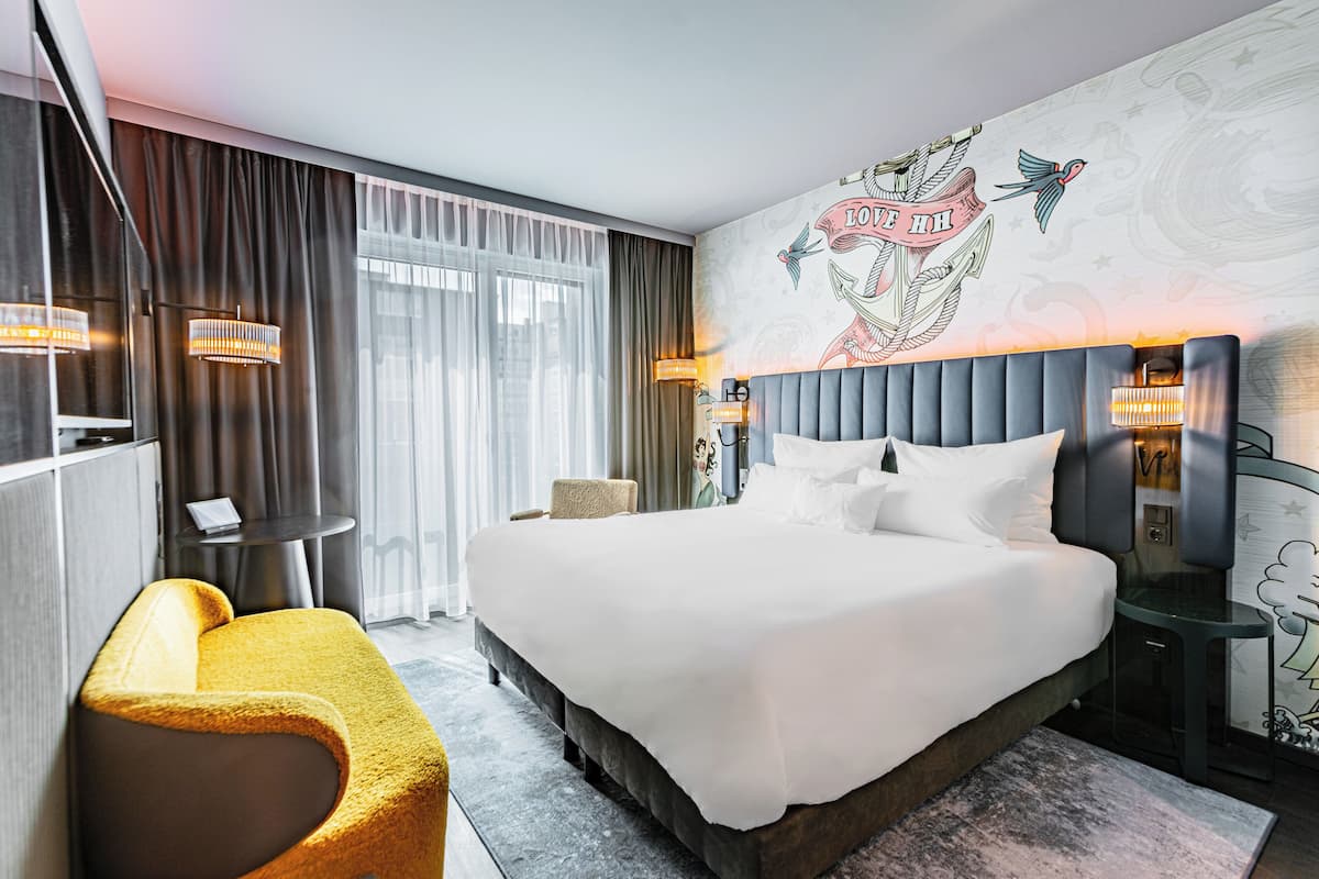 Quarto de hotel moderno com uma cama grande, um sofá, uma mesinha e um mural decorativo na parede.