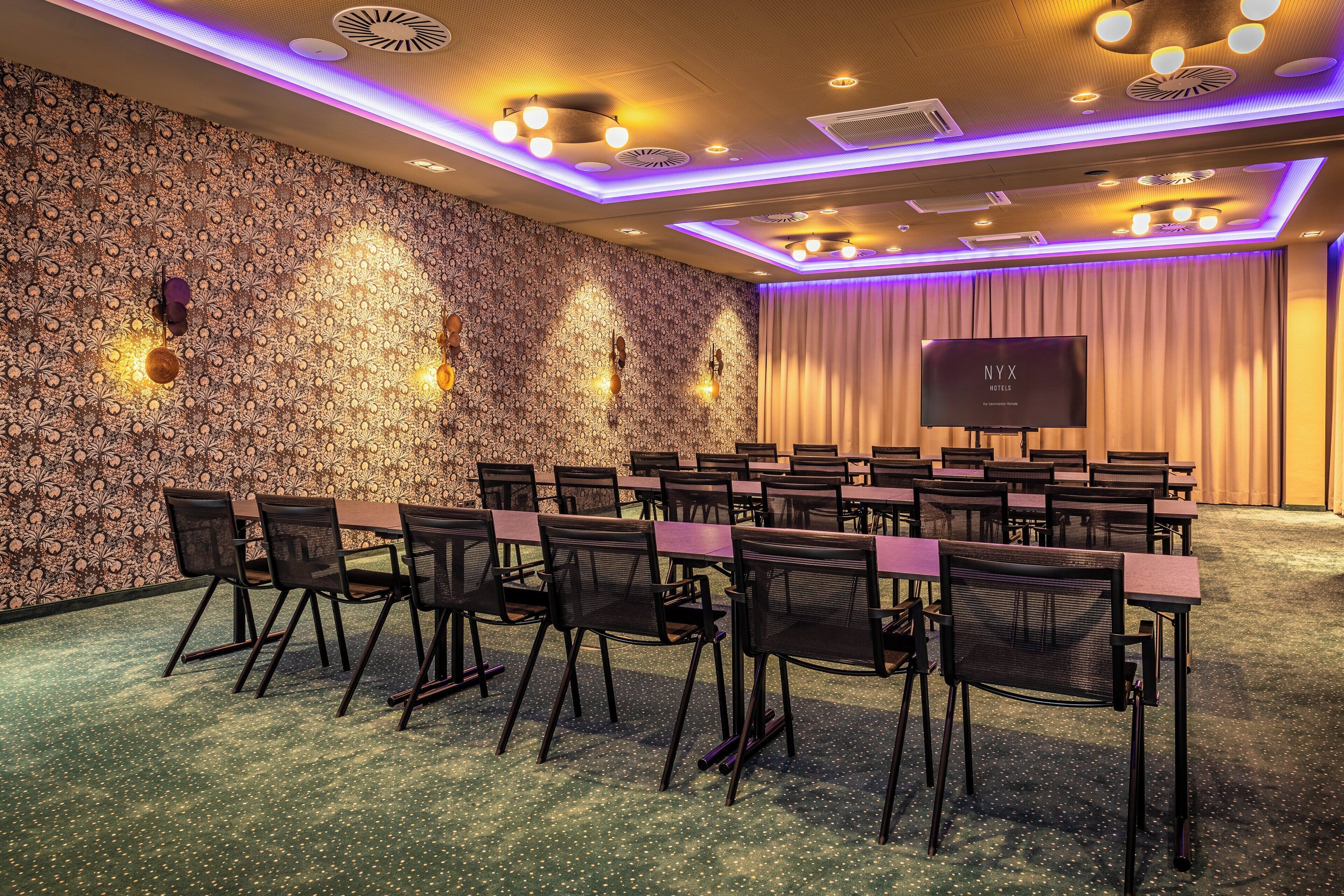 A conference room with a patterned wall, a flat-screen TV, and rows of chairs.