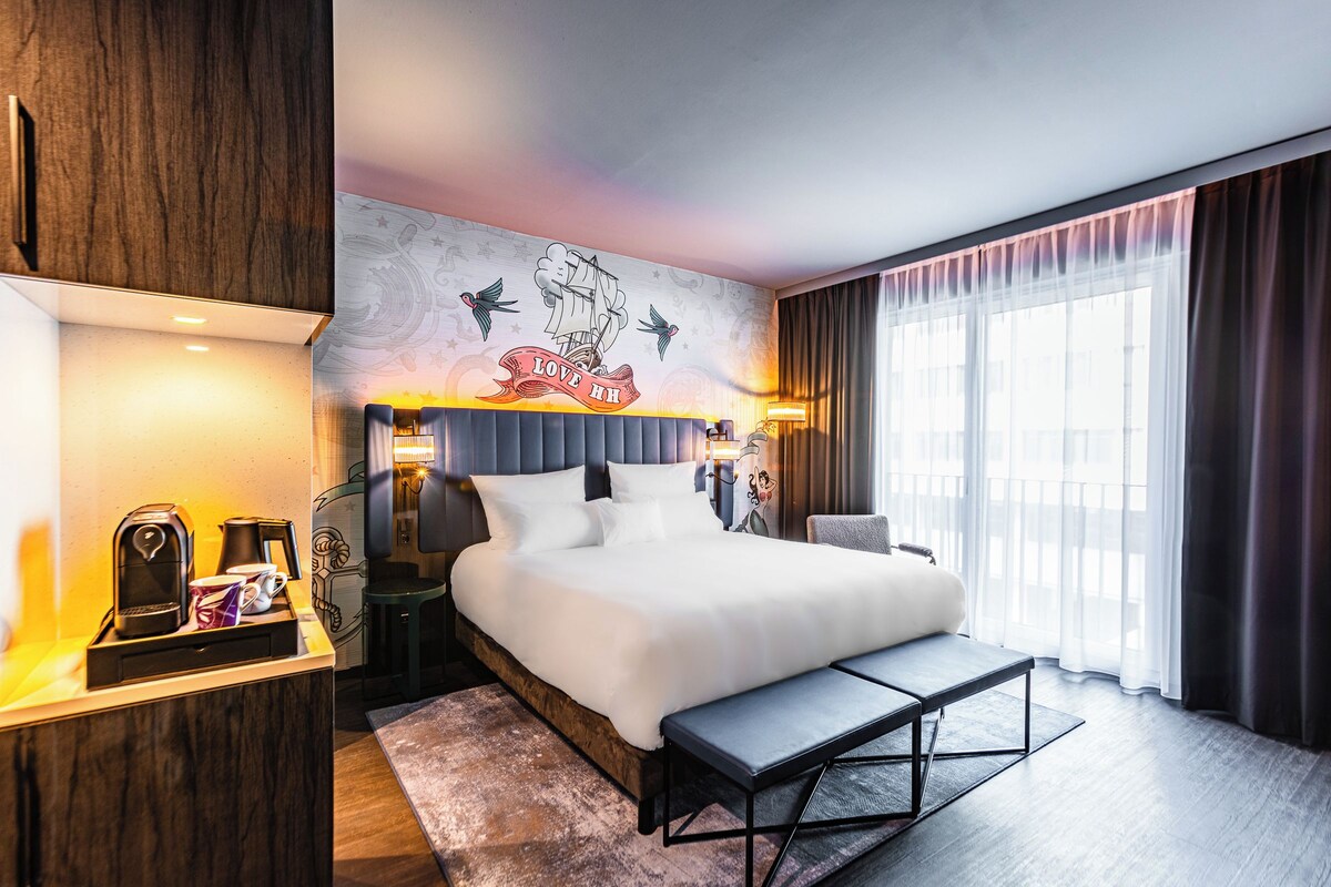 A hotel room with a large bed, bedside tables, a bench, a mural on the wall, and a view of the outdoors.