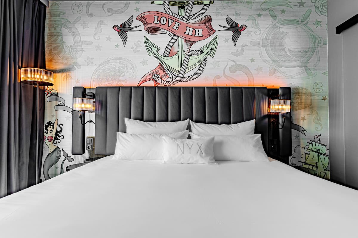 Uma cama bem arrumada com lençóis e almofadas brancas, uma cabeceira estofada escura e uma obra de arte na parede com uma ancora e as palavras "LOVE HH".